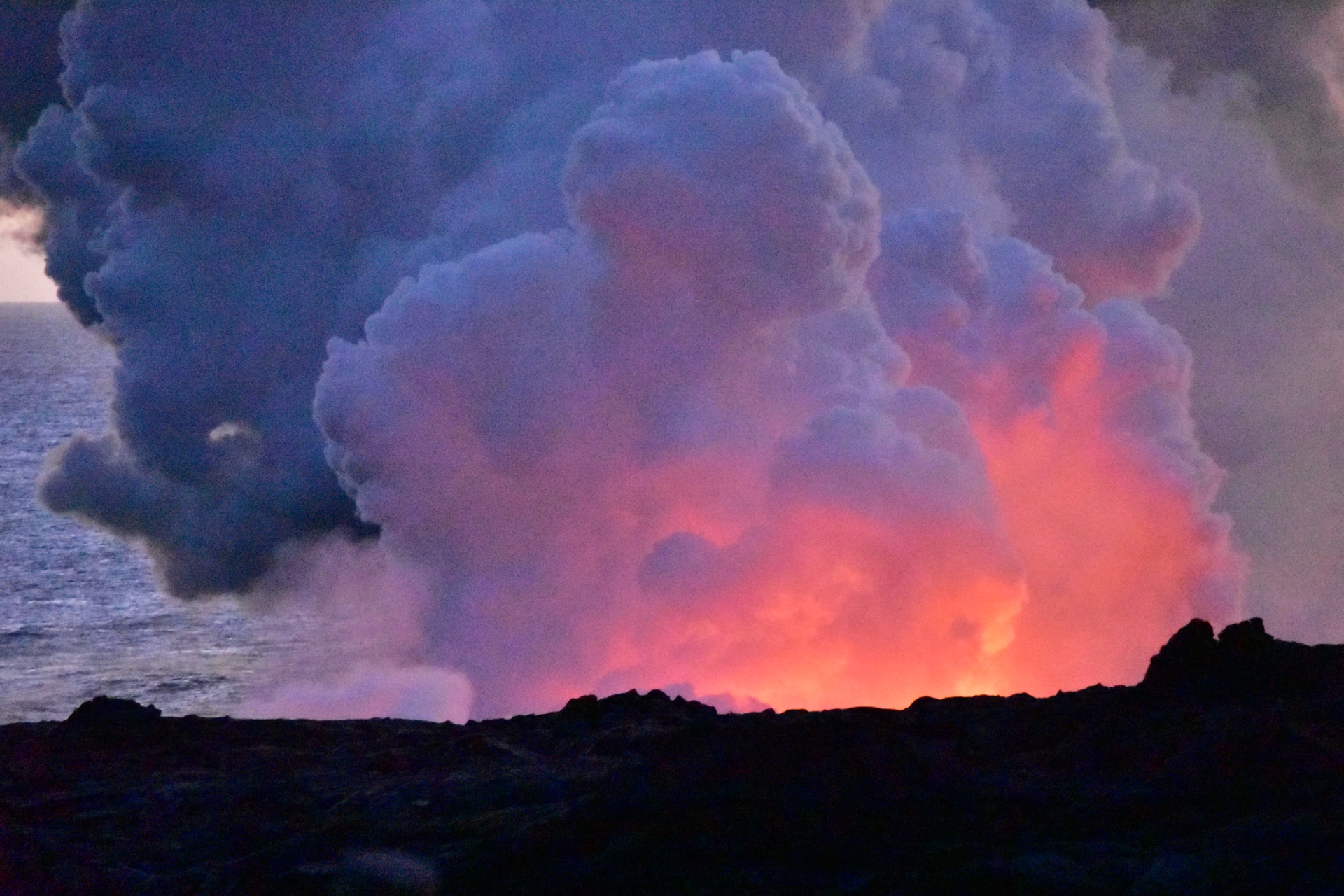 A vast steam plume rising where lava enters the sea, glowing red in the twilight