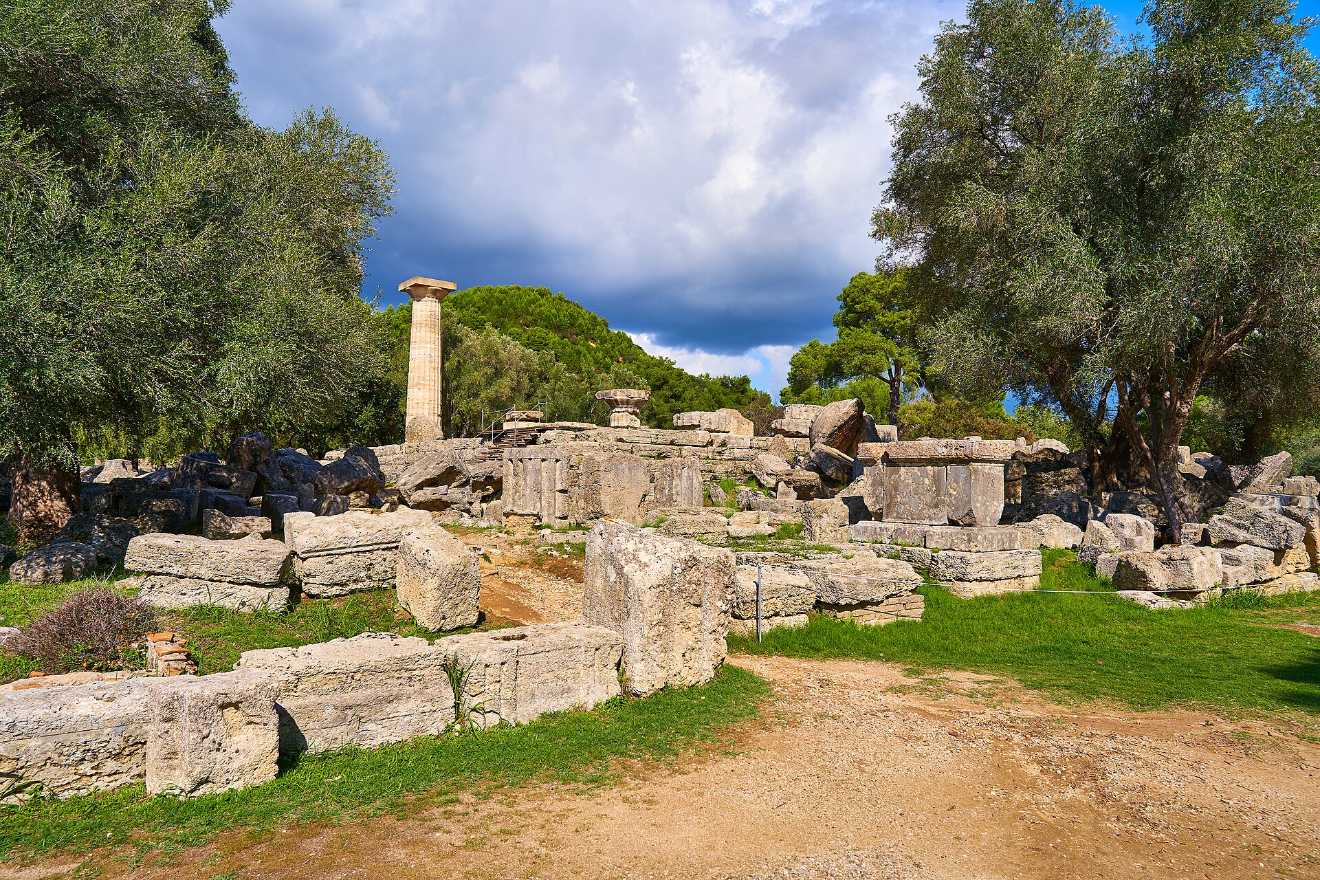 A lone standing column of the Temple of Zeus among olive trees beneath a dramatic sky