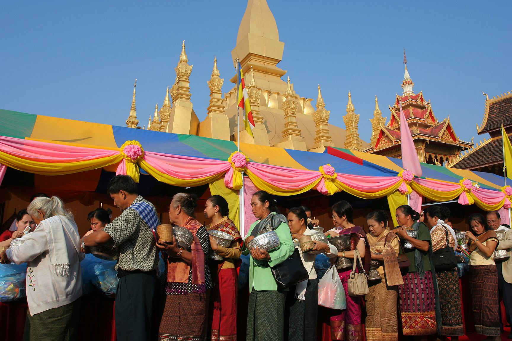 A long line of people holding silver alms bowls before the golden stupa during the That Luang festival