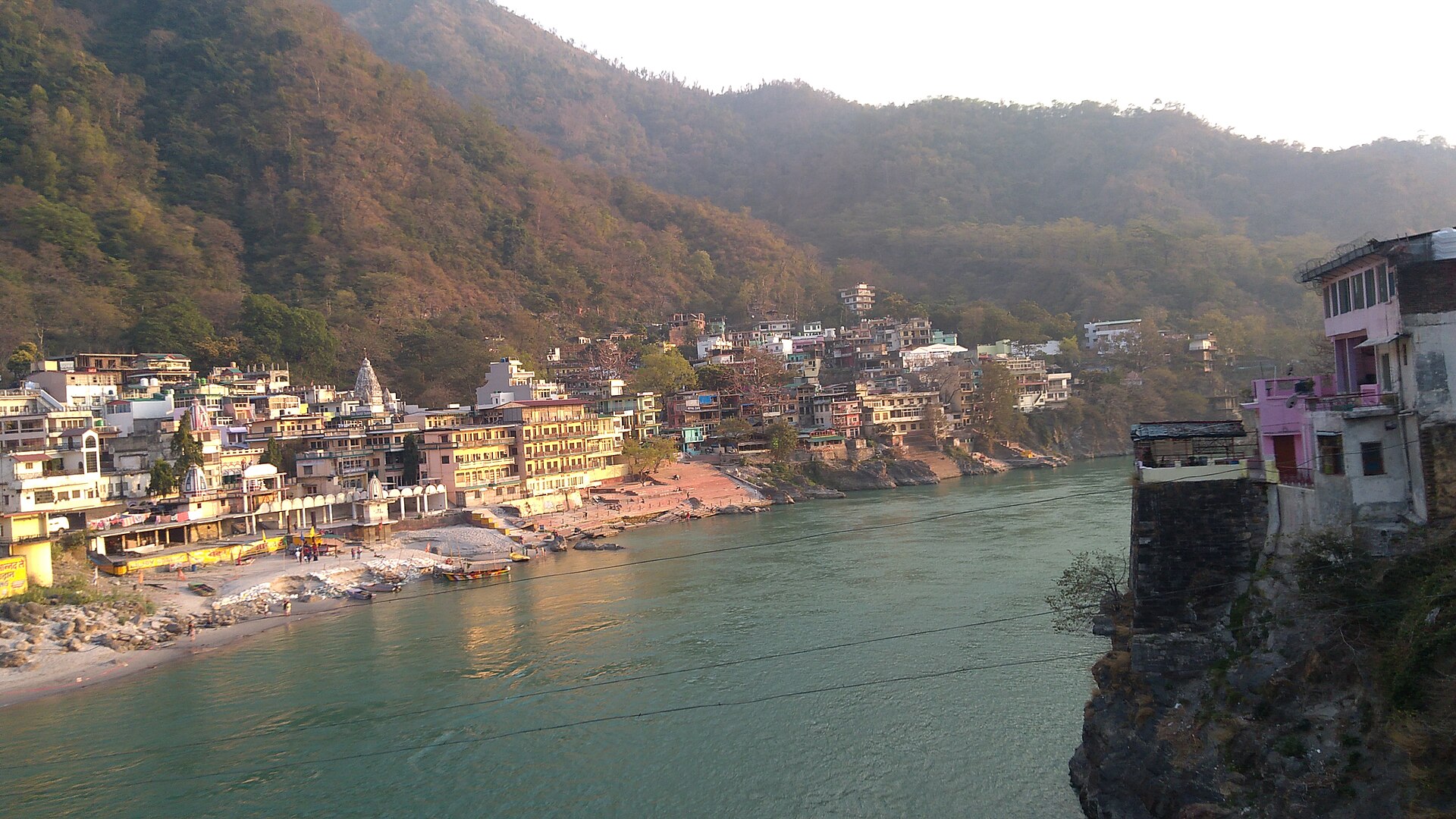 The emerald Ganga flowing between forested hills at dusk, with the riverside ghats of Rishikesh