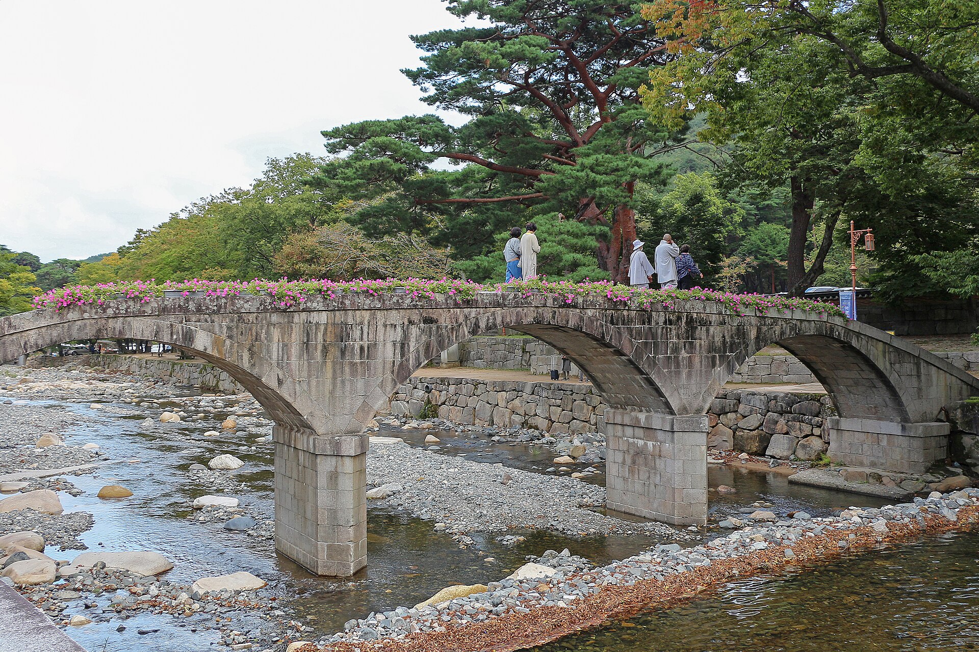 Monks crossing the stone arch bridge over the stream, framed by pines